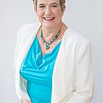 Sue Ritchie standing 3/4 to camera, with a white cardigan and turquoise dress